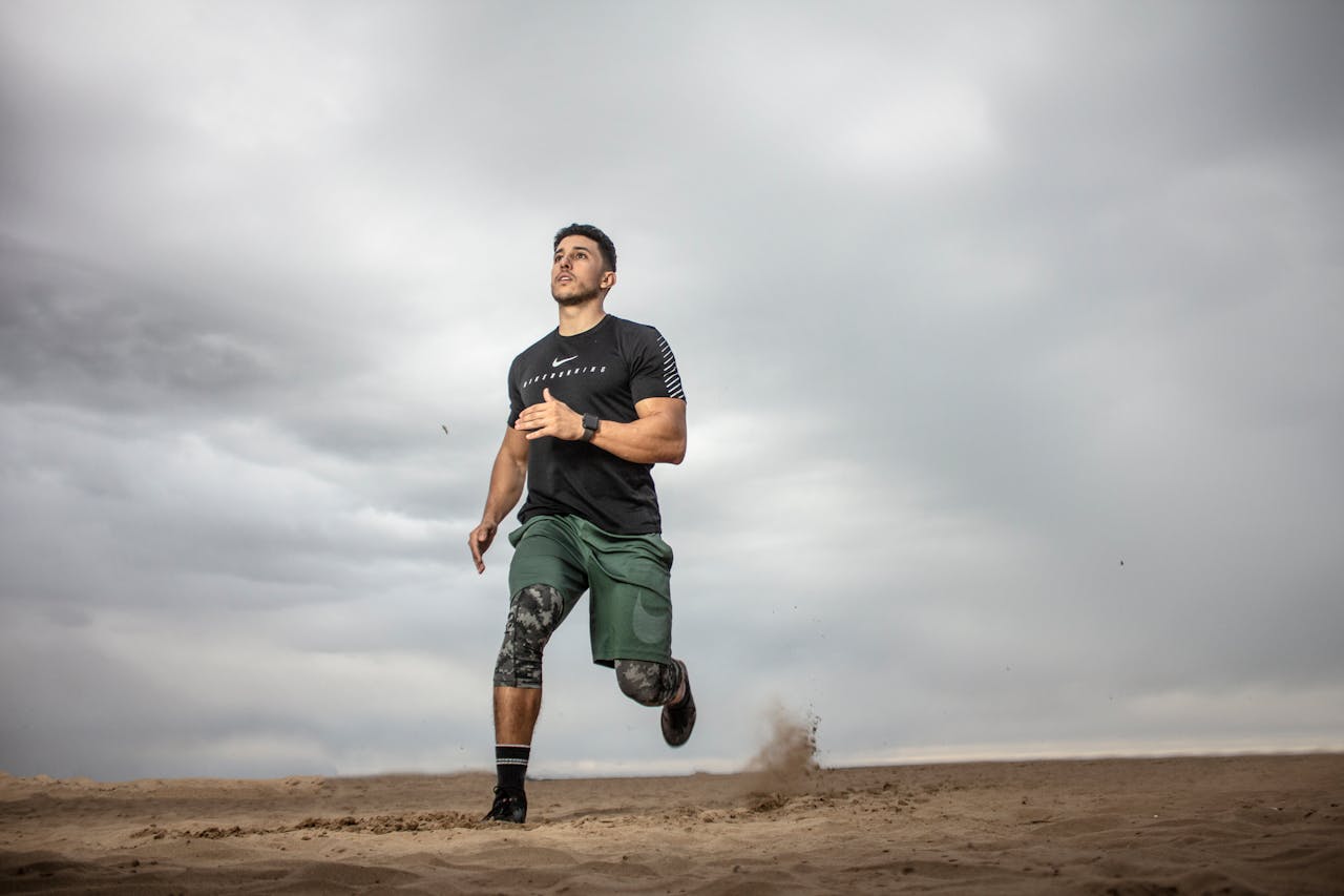 Home A muscular man running on sandy terrain under cloudy skies, promoting fitness and adventure.